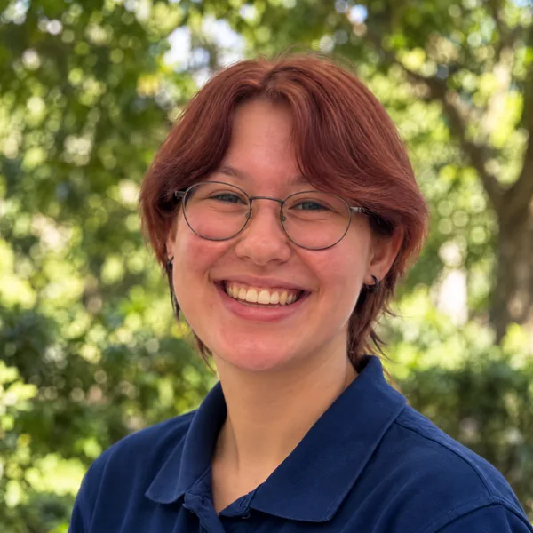 Portrait von Eline mit Brille im blauen Kita-Poloshirt, draussen vor grünen Bäumen